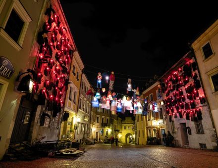 Zdjęcie przedstawia kamienice na Starym Mieście tuż przed Bramą Grodzką w Lublinie. Na dwóch budynkach stojących naprzeciw siebie znajdują się czarne kukły. Ich ciała wykonane są z czarnego, grubego materiału, a ich twarze przypominają wielkie dzioby srok, skierowane w dół. Ich oczy wykonane są z czerwonych, świecących żarówek. Pomiędzy nimi rozwieszony jest sznurek, na którym zawieszone są części kolorowych ubrań. Można tam zobaczyć sukienki w kolorowe geometryczne wzory, niebieskie suknie z falbankami, spódnice w kolorowe groszki oraz zestawy spodni w podobne wzory co suknie i białe koszule. Ubrania te są radosne, jasne i oświetlone białymi żarówkami, co stanowi kontrast wobec ciemnych kukieł na ścianach tuż obok. Wokół panuje mrok, a lampy na instalacji stanowią dodatkowe źródło oświetlenia ulicy, razem z lampami wiszącymi na kilku budynkach bliżej Bramy Grodzkiej. Zdjęcie przedstawia kamienice na Starym Mieście tuż przed Bramą Grodzką w Lublinie. Na dwóch budynkach stojących naprzeciw siebie znajdują się czarne kukły. Ich ciała wykonane są z czarnego, grubego materiału, a ich twarze przypominają wielkie dzioby srok, skierowane w dół. Ich oczy wykonane są z czerwonych, świecących żarówek. Pomiędzy nimi rozwieszony jest sznurek, na którym zawieszone są części kolorowych ubrań. Można tam zobaczyć sukienki w kolorowe geometryczne wzory, niebieskie suknie z falbankami, spódnice w kolorowe groszki oraz zestawy spodni w podobne wzory co suknie i białe koszule. Ubrania te są radosne, jasne i oświetlone białymi żarówkami, co stanowi kontrast wobec ciemnych kukieł na ścianach tuż obok. Wokół panuje mrok, a lampy na instalacji stanowią dodatkowe źródło oświetlenia ulicy, razem z lampami wiszącymi na kilku budynkach bliżej Bramy Grodzkiej.