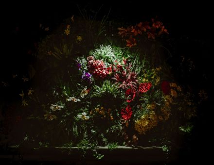 Zdjęcie przedstawia konstrukcje roślinną. Jest to zielona konstrukcja z różnego rodzaju gęsto ułożonymi roślinami. W niektórych miejscach są włożone czerwone duże kwiaty. Niektóre są fioletowe, inne białe i pomarańczowe, które luźno zwisają po liściach. Wokół konstrukcji jest ciemno i tylko jej środek jest oświetlany białym światłem. Zdjęcie przedstawia konstrukcje roślinną. Jest to zielona konstrukcja z różnego rodzaju gęsto ułożonymi roślinami. W niektórych miejscach są włożone czerwone duże kwiaty. Niektóre są fioletowe, inne białe i pomarańczowe, które luźno zwisają po liściach. Wokół konstrukcji jest ciemno i tylko jej środek jest oświetlany białym światłem.