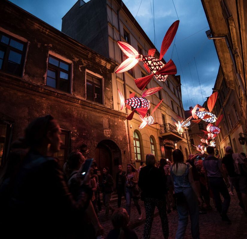 Zdjęcie zrobione nocą. Ludzie przyglądają się instalacji artystycznej podświetlanym rybom wiszącym nad ich głowami.